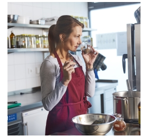 In einer Küche steht eine Frau mit einer Schürze, die an einem Gewürz riecht. 